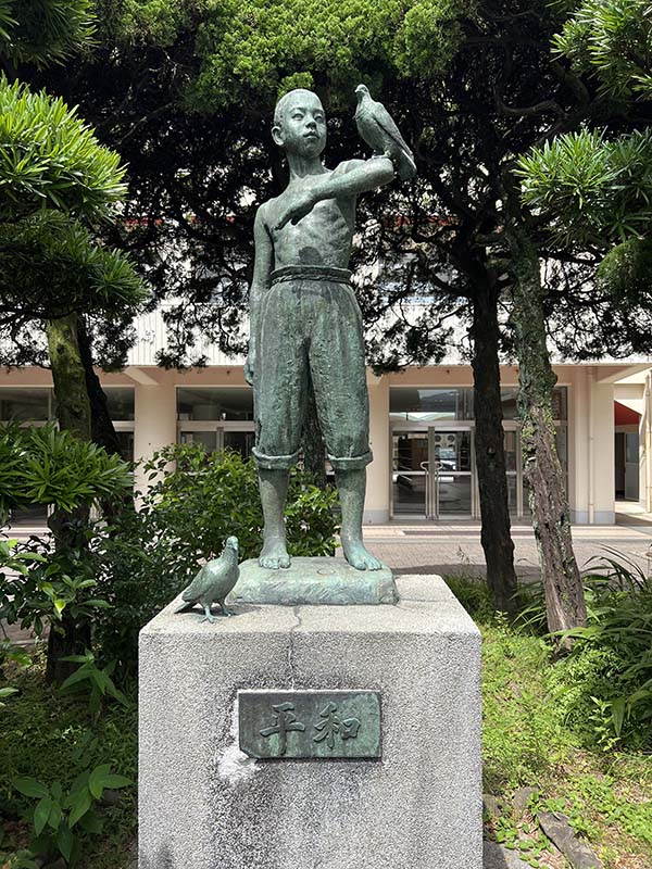 Statue of a boy wearing rolled up trousers with a bird on his arm and a bird at his feet. The statue is on a plinth with trees in the background.
