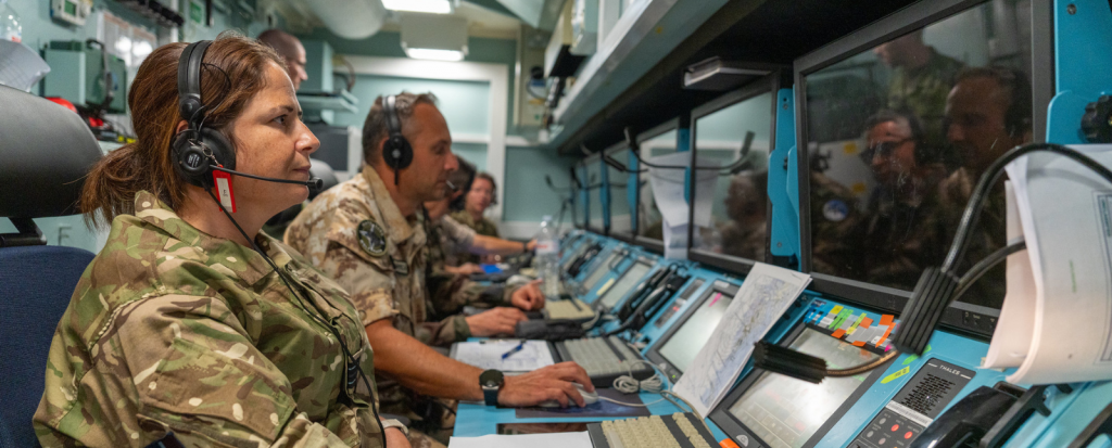 Military personnel in a control room wearing headsets and working at computer consoles with multiple screens.