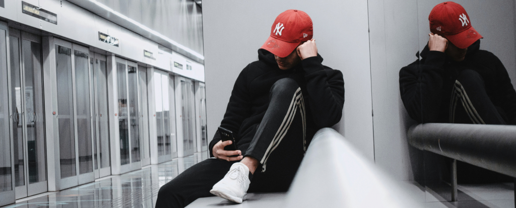 Young person in a red cap, black hoodie and white trainers sits on a bench, looking down at their phone. Behind them are automatic station doors.