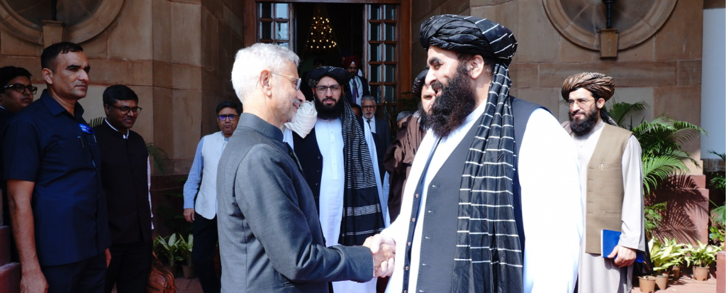India External Affairs Minister Dr. S. Jaishankar and Minister of Foreign Affairs of Afghanistan, Amir Khan Muttaqi shake hands outside a building. Jaishankar wears a grey suit, Muttaqi a black turban and waistcoat over a white shirt with striped scarf.