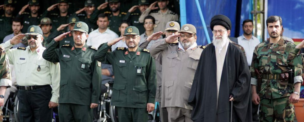 Ali Khamenei has a white beard and glasses. He stands in front of men in army gear who are saluting. Source: Khamenei.ir. Wikimedia Commons.CC BY-SA 4.0